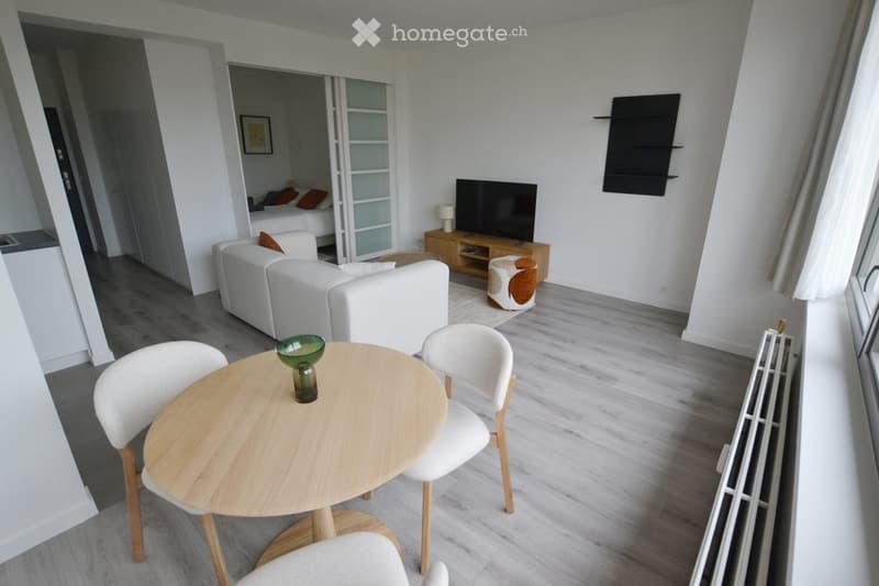 Modern living room with white sofa, small round wooden table, white chairs, television on wooden stand, and a view of a bedroom through an open doorway.