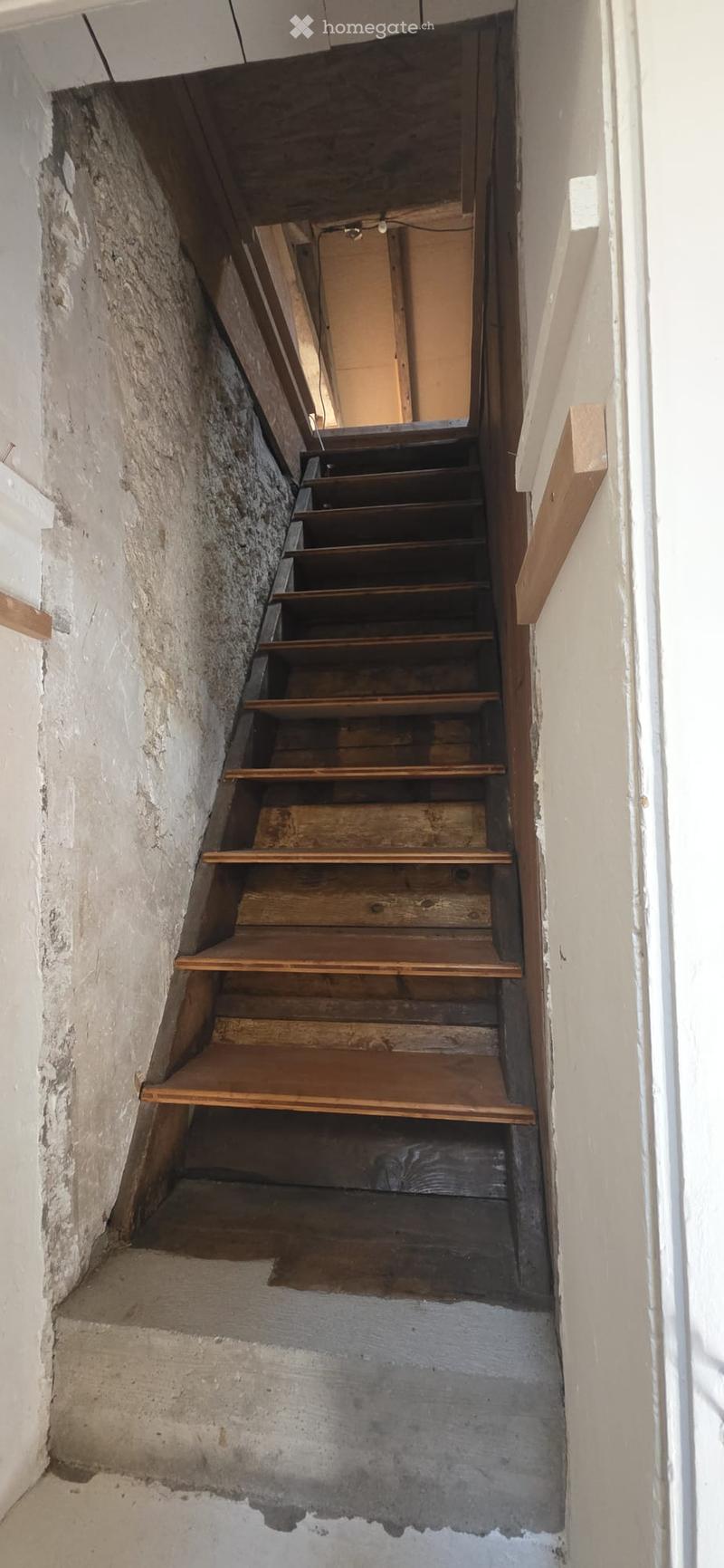 A narrow, steep staircase with wooden steps and a metal railing, leading upwards. The walls are unfinished with visible plaster and some wear.