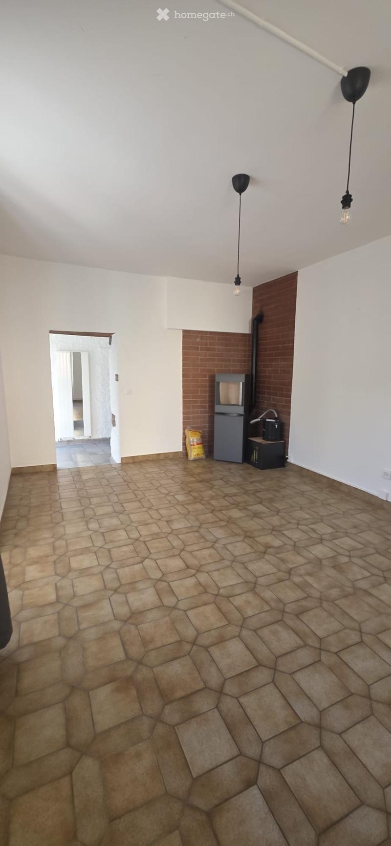 Empty room with hexagonal tiled floor, white walls, and exposed brick section. Minimal lighting with two pendant lamps.