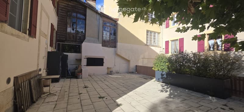 Courtyard with tiled pavement, small table, and surrounding buildings with shutters and windows.