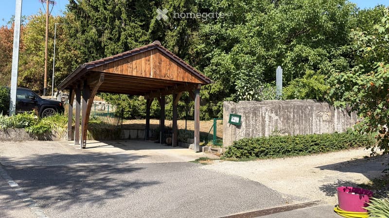 A wooden carport with a slanted roof is situated on a gravel driveway, surrounded by greenery and a stone wall. A black SUV is parked nearby, and a pink trash bin is visible on the right side.