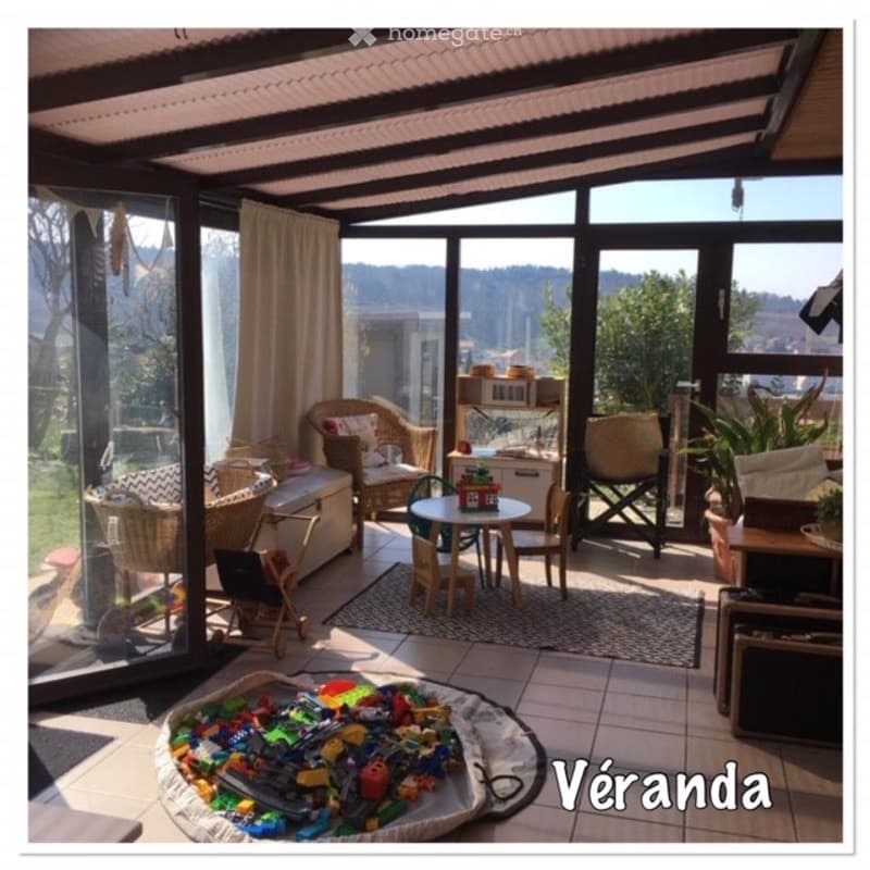 A sunlit enclosed patio with large glass walls, featuring a sofa, small dining table with chairs, storage unit, and a rug with toys on the floor.