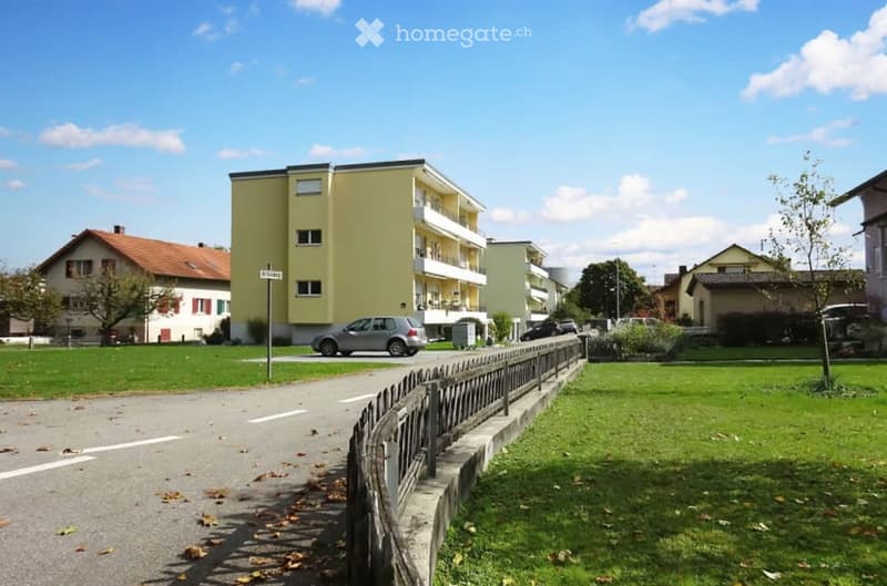 Yellow apartment building, several parked cars, small pathway, road, grass, trees