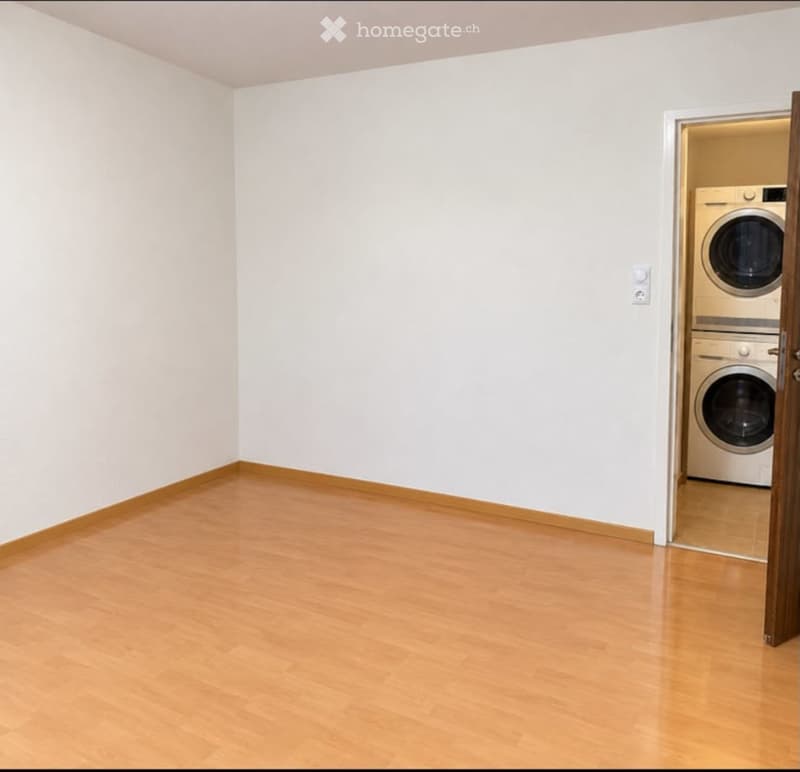 Empty room with light wooden flooring and white walls, featuring an open doorway leading to a private washing machine area.