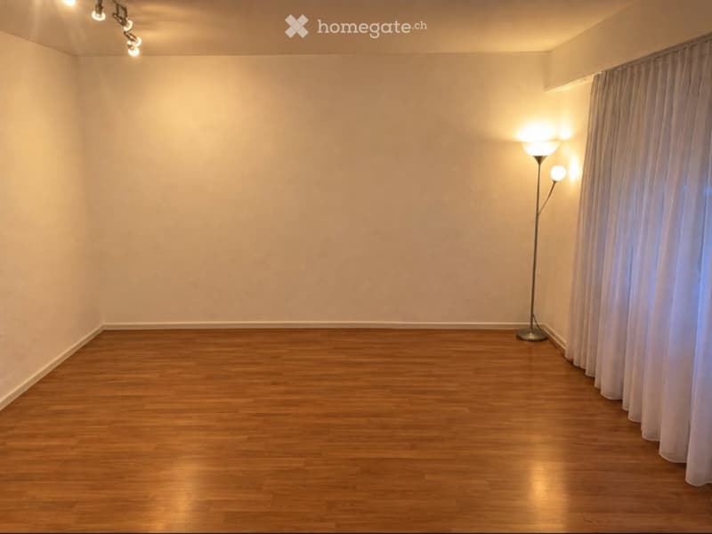 Empty room with wooden flooring, white walls, and a single floor lamp near a window with curtains.