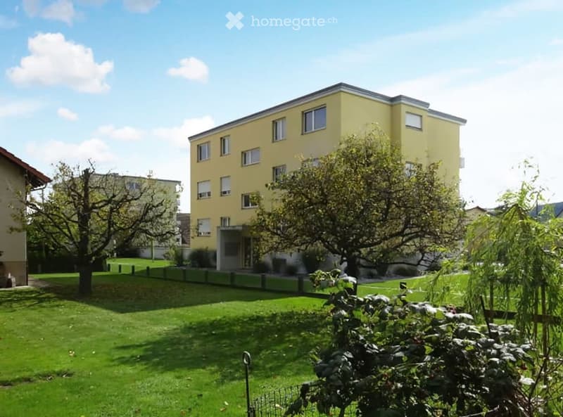 Apartment building with a yellow facade, multiple windows, fenced lawn, and trees in the yard