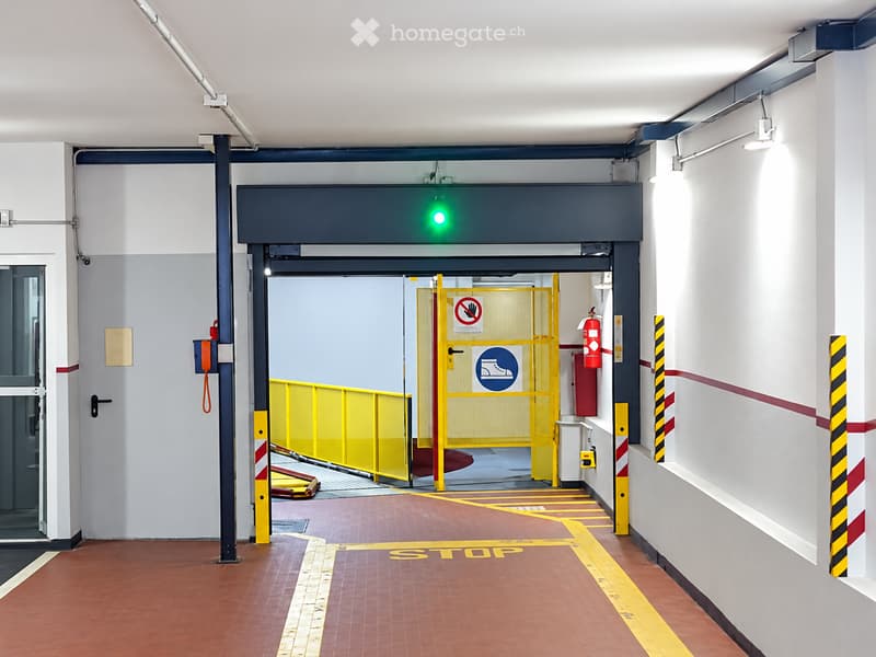 A modern, clean corridor leading to a parking area with a yellow barrier gate, red floor tiles, and safety signage.