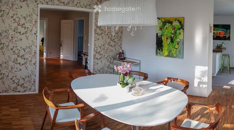 Dining room with a round white table, six wooden chairs, a vase with pink flowers, and a painting on the wall. The room features wooden flooring and a chandelier hanging from the ceiling.