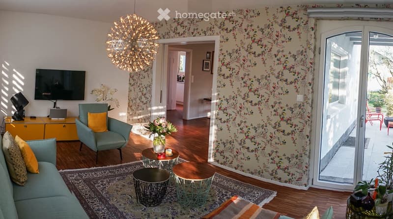 Modern living room with a floral wallpaper accent wall, a teal sofa with yellow pillows, a matching armchair, a coffee table, side tables, a television, and a decorative flower vase on a side table. The room features a patterned rug and a hanging light fi