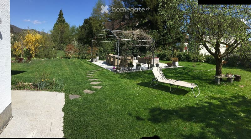 Well-maintained garden with a lounge area, sun lounger, and dining set under a pergola, surrounded by trees and greenery.