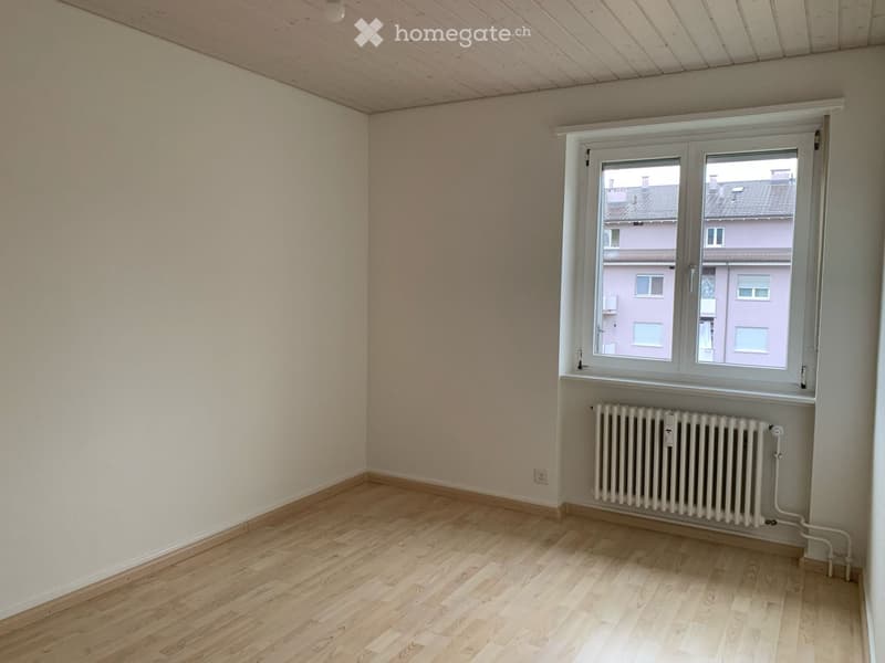 Empty room with light wooden flooring, white walls, and a single window with a radiator below it.