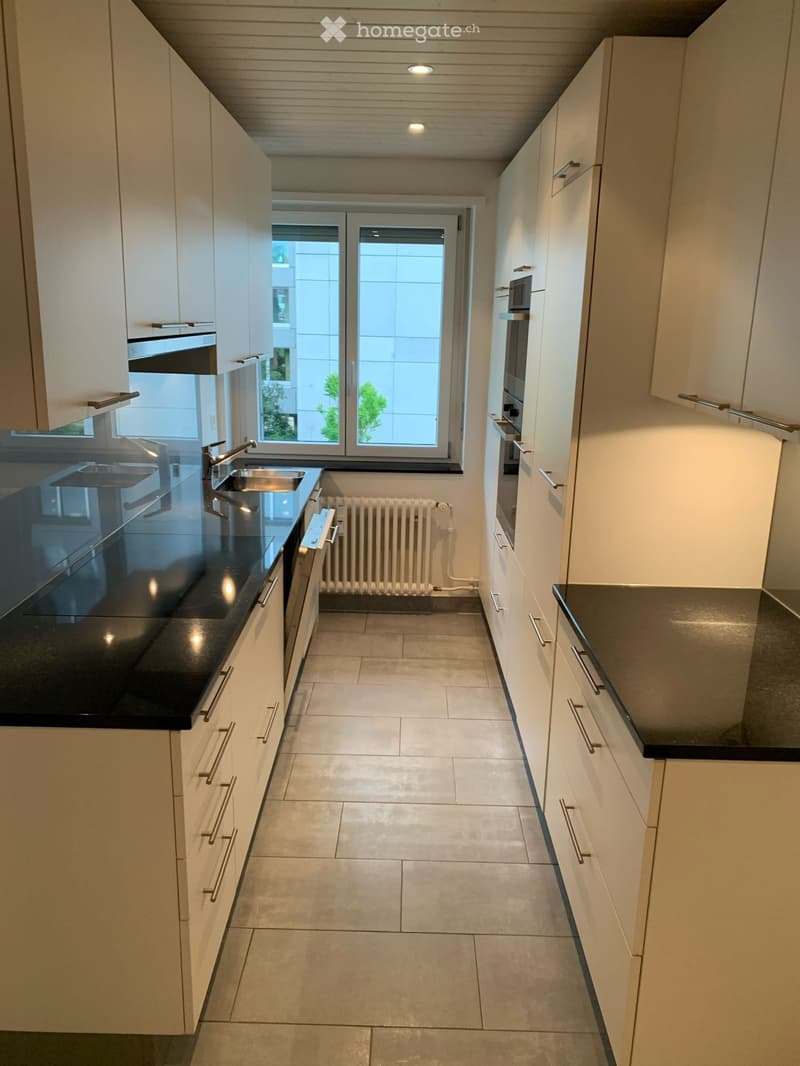 Modern kitchen with white cabinets, black countertops, a sink, an oven, and a radiator. The space is well-lit with natural light from a large window.