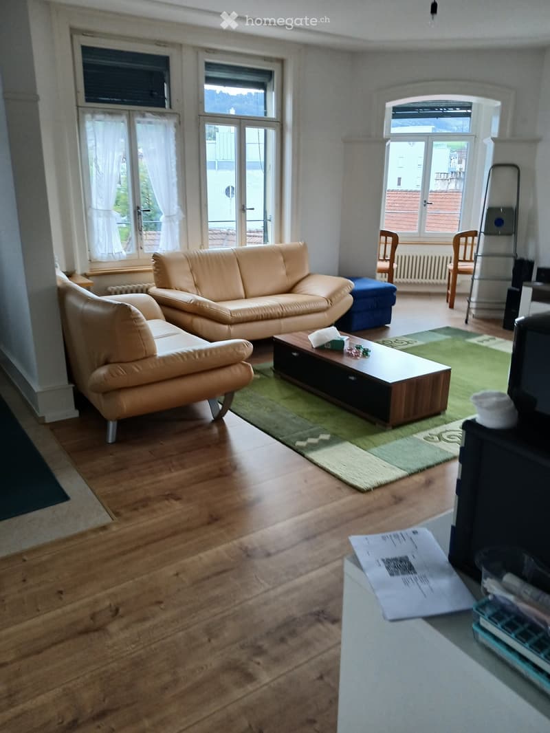Living room with light-colored leather sofa, coffee table, chairs, and a fireplace. The room features wooden flooring and large windows with white curtains.