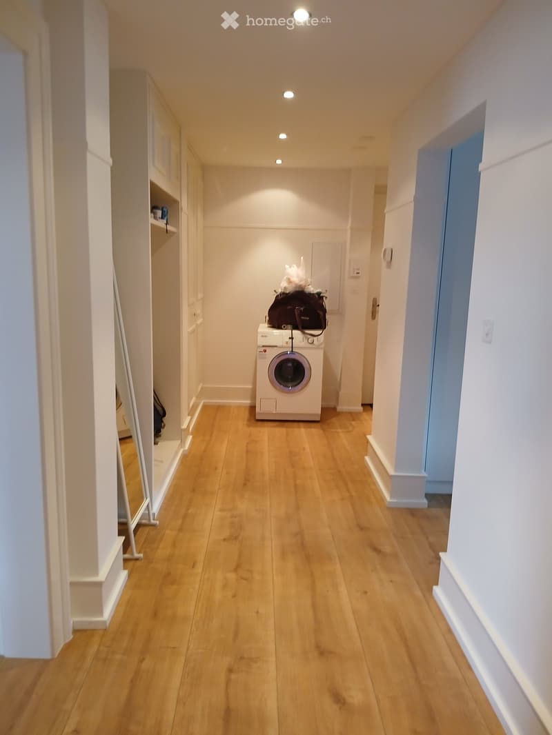 A narrow hallway with wooden flooring, white walls, and recessed lighting. A washing machine is placed in the center, with a bag on top. Built-in cabinets line the left wall.