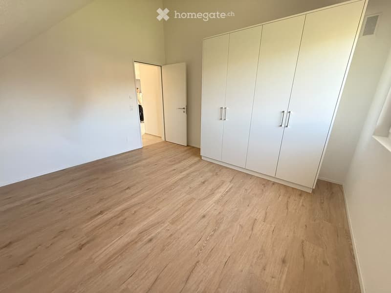 Empty room with light wooden flooring, white walls, and built-in white wardrobes. A doorway leads to another room.