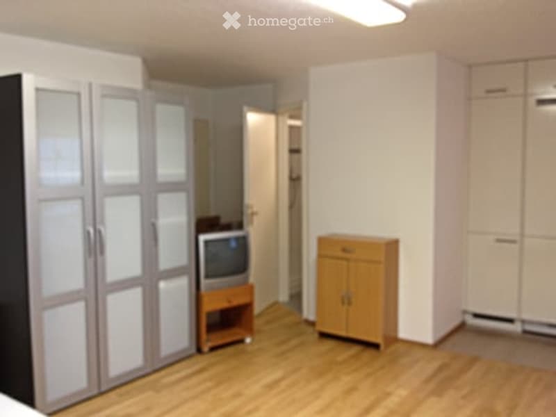 A small, furnished room with light wooden flooring, white walls, and a large mirrored wardrobe. A small TV sits on a wooden stand, and a wooden cabinet is placed near the door.