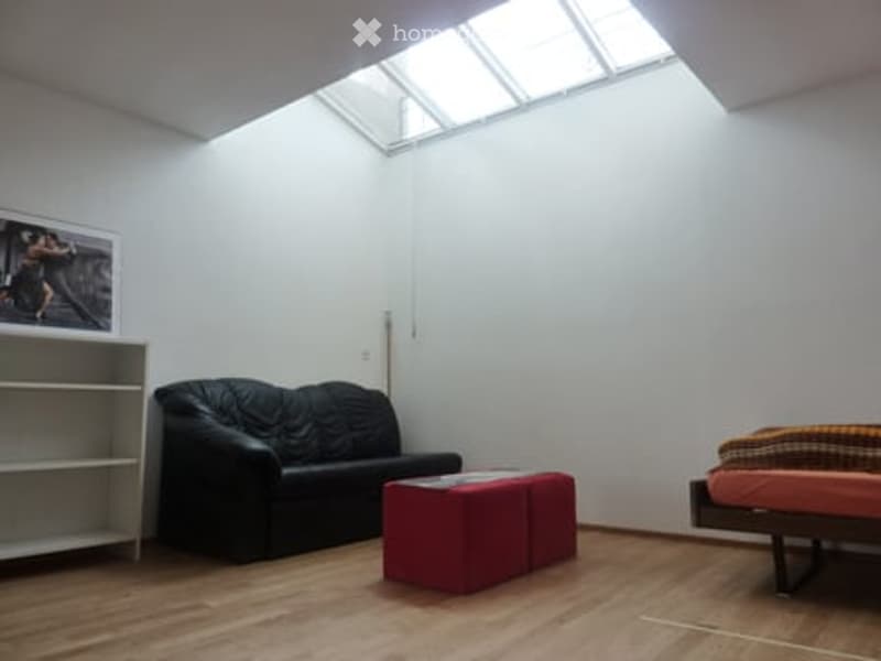A modern, minimalist room with a black sofa, red ottoman, bed with colorful bedding, a white shelf with a picture frame, and wooden flooring.