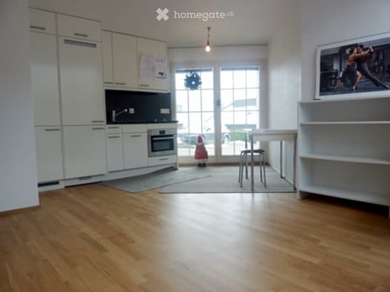 Bright, modern kitchen with white cabinets, an oven, a small table with a stool, and a large screen on the wall. The floor is made of light wood, and there is a small rug in front of the table.