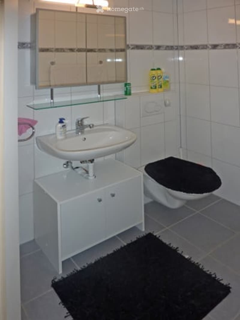 Modern bathroom with white tiles, a white sink, a toilet with a black seat cover, a mirror above the sink, and a black rug on the floor.