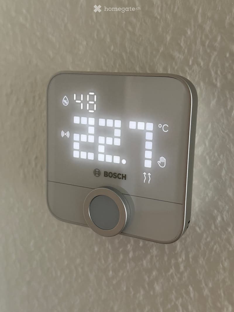 A Bosch thermostat mounted on a wall, displaying temperature and humidity levels with a digital interface and control knob.
