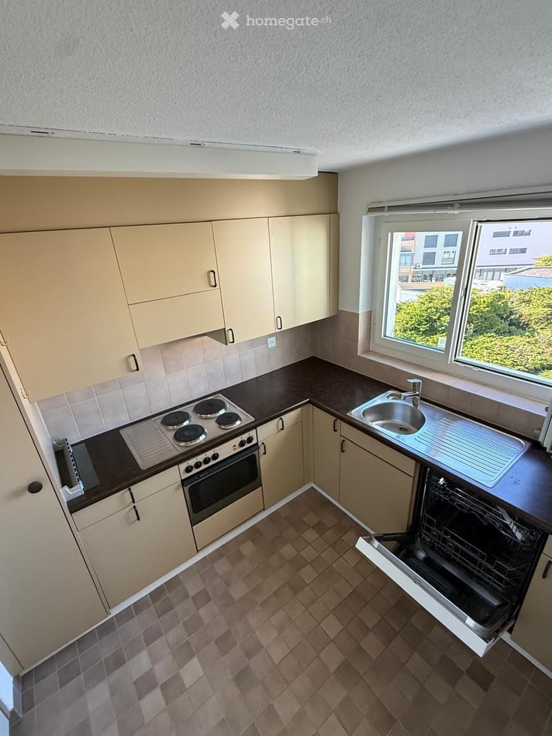 Compact kitchen with light-colored cabinets, a four-burner electric stove, a sink, and an open dishwasher. The space features tiled walls and a tiled floor.