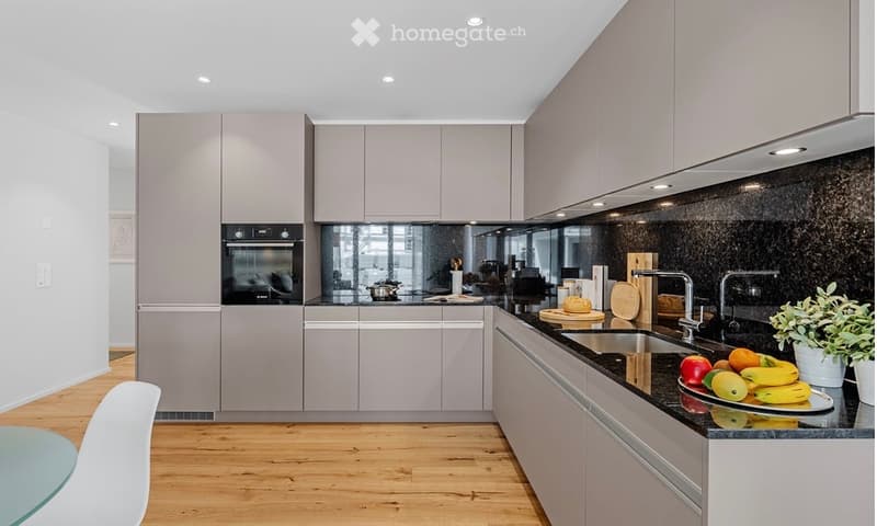 Modern kitchen with light grey cabinets, black countertops, stainless steel appliances, and a wooden floor. Features include an oven, sink, faucet, cutting board, bowl, and a small table with a chair.
