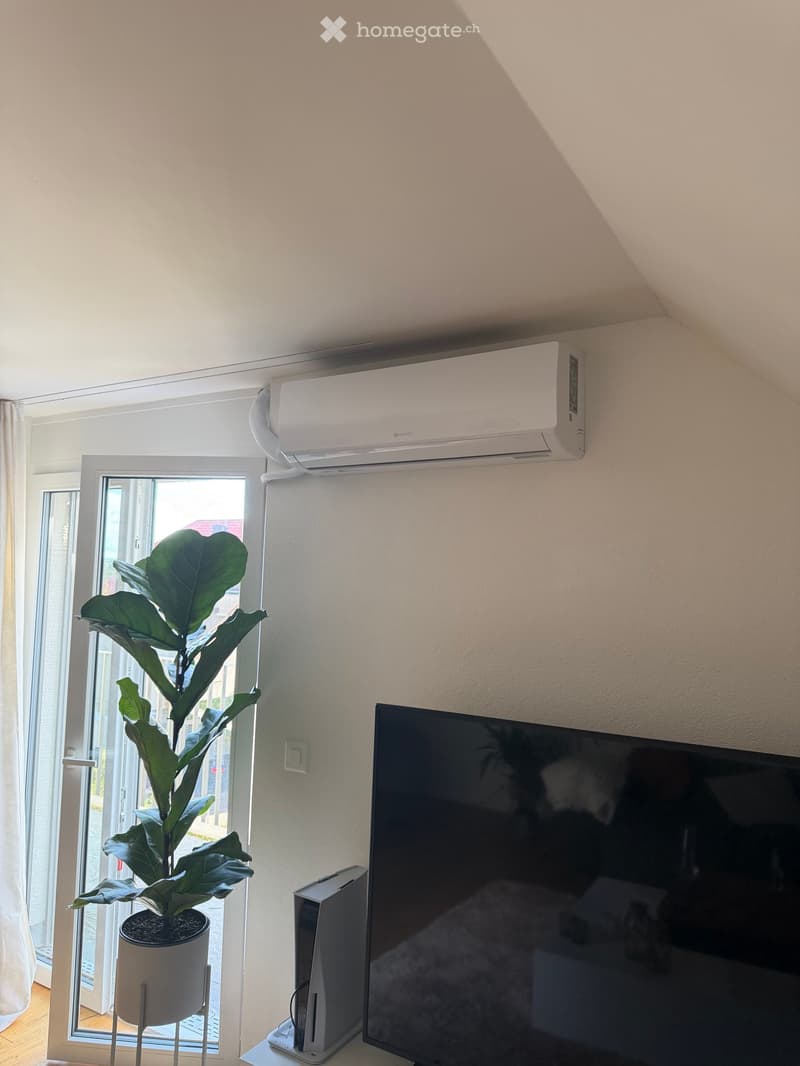 A modern living room with a large window, a potted plant, a television, and an air conditioner mounted on the wall.