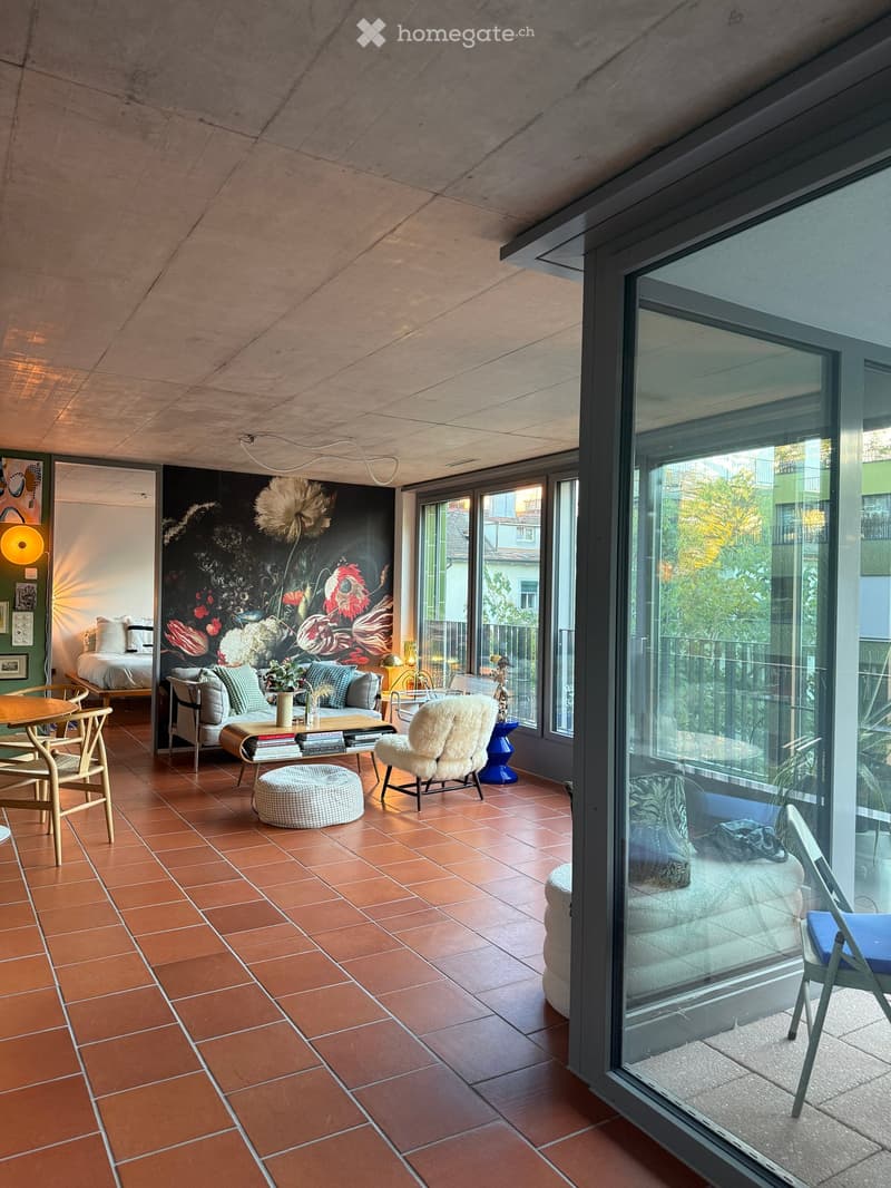 Modern furnished room with terracotta tiles, large windows, sofa, coffee table, ottoman, chair, and bed visible.