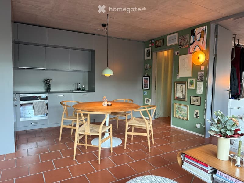 Open-plan kitchen and dining area with grey cabinets, a round wooden dining table with four chairs, a pendant light, and a vase with flowers on a side table.