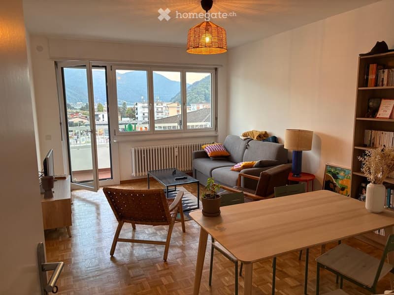 Modern living room with large windows offering a scenic mountain view, featuring a grey sofa, coffee table, wooden dining table, wicker chair, and decorative lamps.