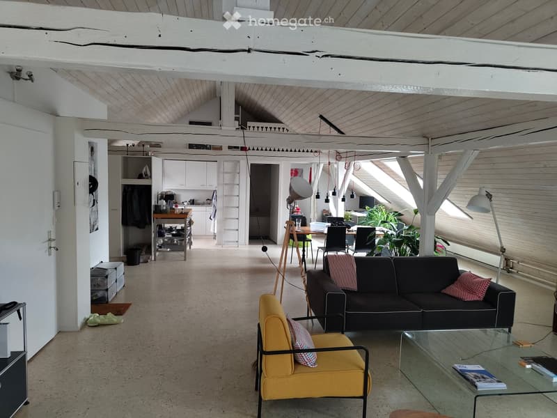 Open-plan living and dining area with exposed wooden beams, concrete floor, and large windows. Features a black sofa, yellow armchair, glass coffee table, and a small kitchen area with white cabinets and a wooden stool.