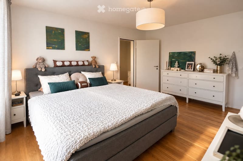 Cozy bedroom with a grey upholstered bed, white bedding, decorative pillows, a nightstand with a lamp, a dresser with drawers, and artwork on the walls.