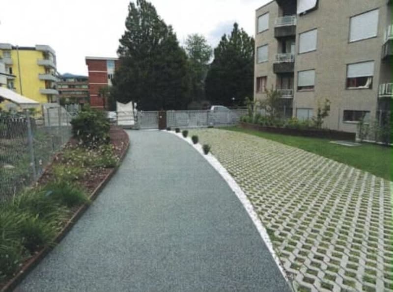 A paved pathway leads to a gated entrance, flanked by small plants and shrubs, with a multi-story apartment building visible in the background.