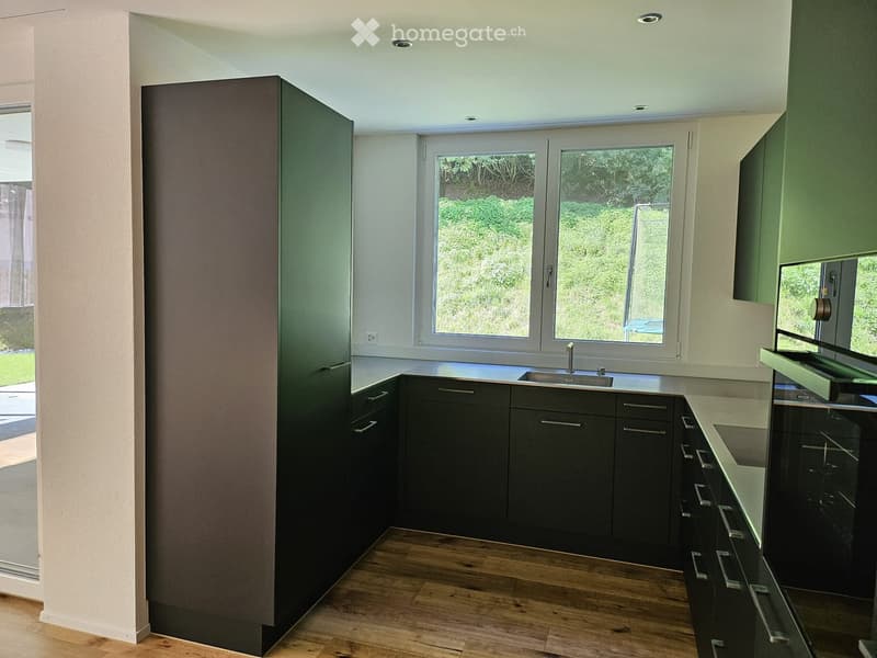 Modern kitchen with dark grey cabinets, a stainless steel sink, and a wooden floor. The room features large windows with a view of greenery outside.