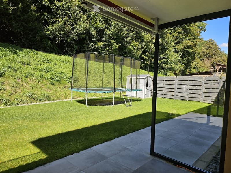 Garden with a trampoline, wooden shed, and wooden fence, surrounded by lush greenery and trees.