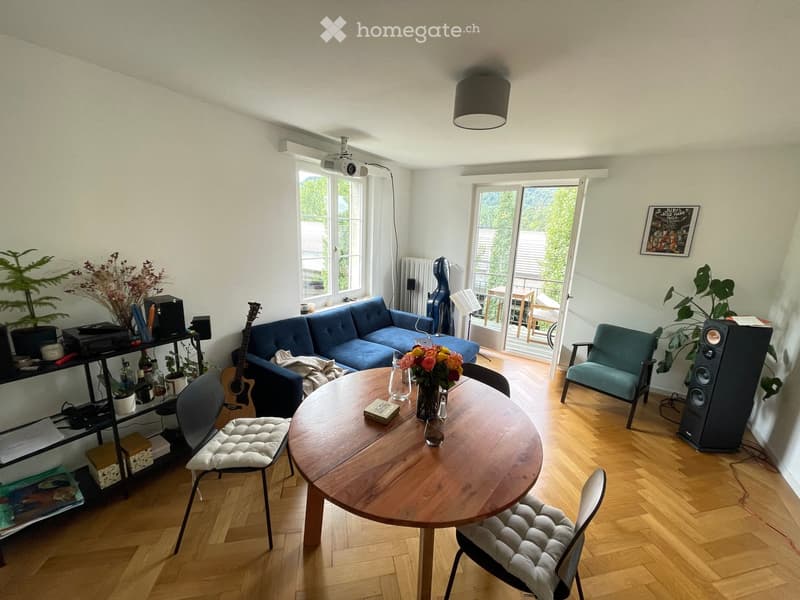 Living room with wooden floor, large windows, blue sofa, round wooden table, chairs, guitar, speaker, plants, and a view of the balcony.