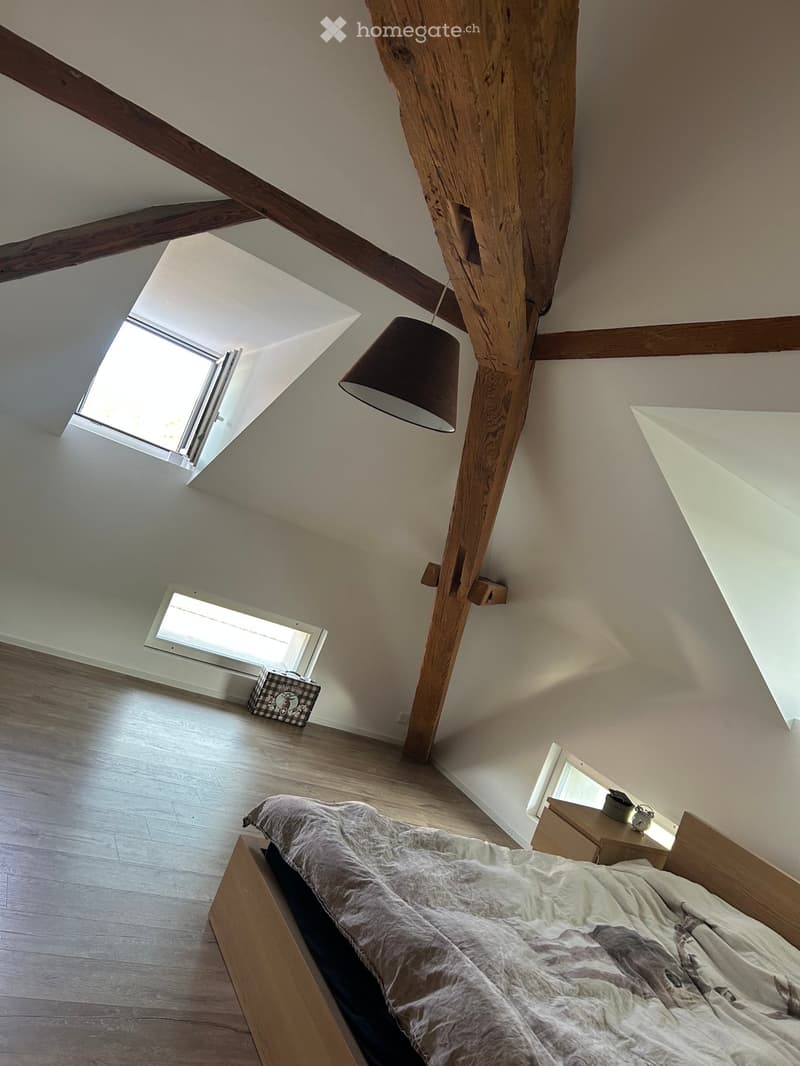 Cozy attic bedroom with wooden beams, skylight windows, a wooden bed with a textured duvet, a nightstand with a lamp, and a small suitcase in the corner.