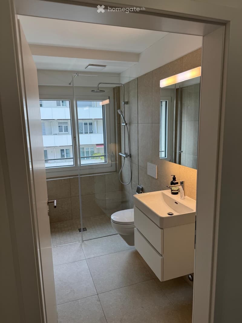 Modern bathroom with a glass-enclosed shower, a white sink with a built-in cabinet, a toilet, and a large mirror with built-in lighting. The space features tiled walls and floors and has a window offering a view of the exterior.