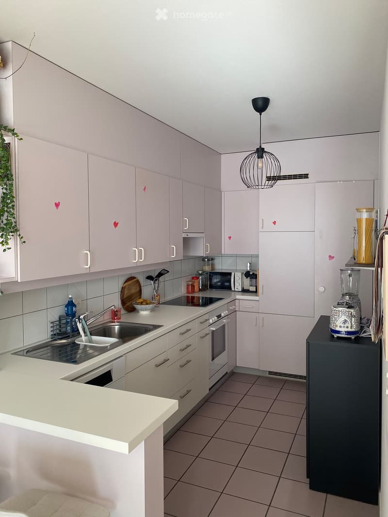 Modern kitchen with light grey cabinets, white countertops, stainless steel sink, and various appliances including a microwave, oven, blender, and toaster. Features a hanging pendant light and a small island with a sink.