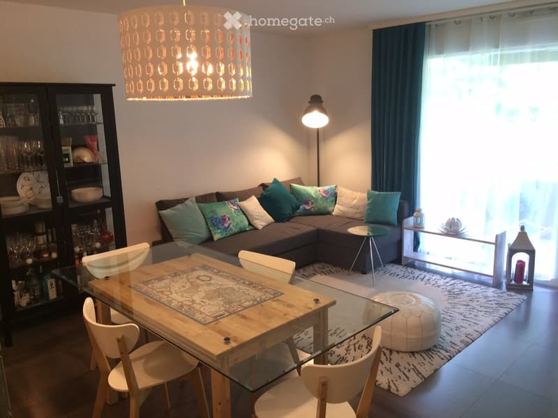 Modern living room with a brown sectional sofa, colorful cushions, a glass coffee table, a wooden dining table with chairs, a white ottoman, and a black cabinet with glassware.