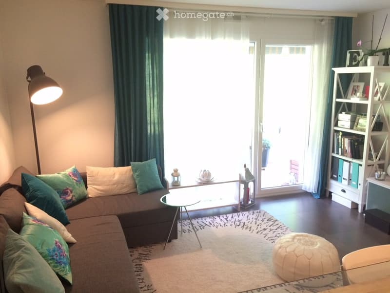 Modern living room with a brown sectional sofa, teal throw pillows, a glass coffee table, a white ottoman, a white shelving unit, and a floor lamp near a large window with green curtains.