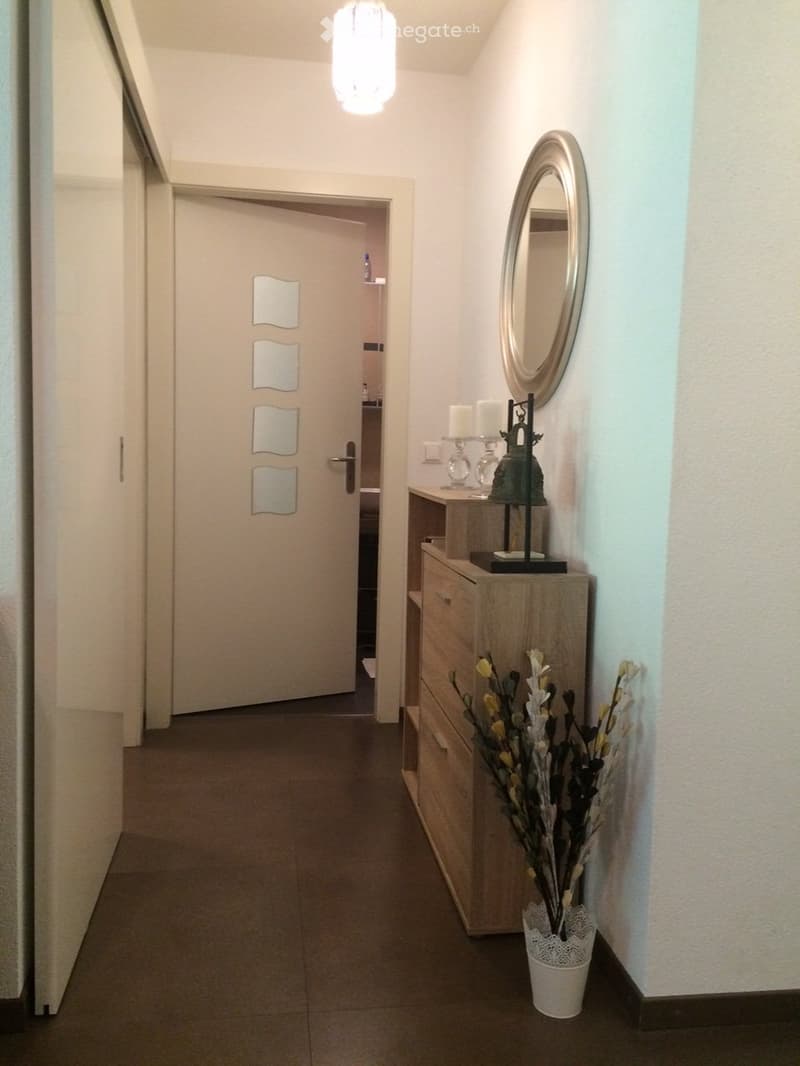 A clean, modern hallway with a tiled floor, white walls, a ceiling light fixture, a small wooden console table with a decorative vase, and a mirror on the wall.