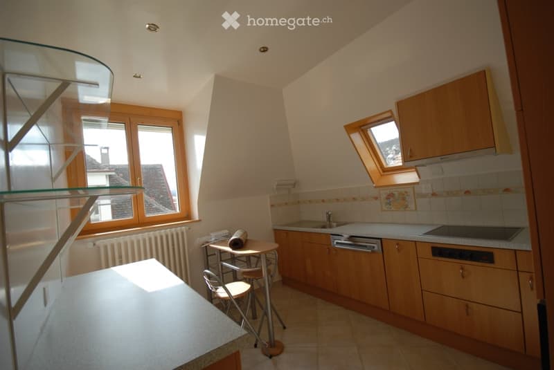 A small, well-lit kitchen with wooden cabinets, a sink, a dishwasher, and a small dining area with a table and chairs. The room features large windows and a skylight, allowing ample natural light.