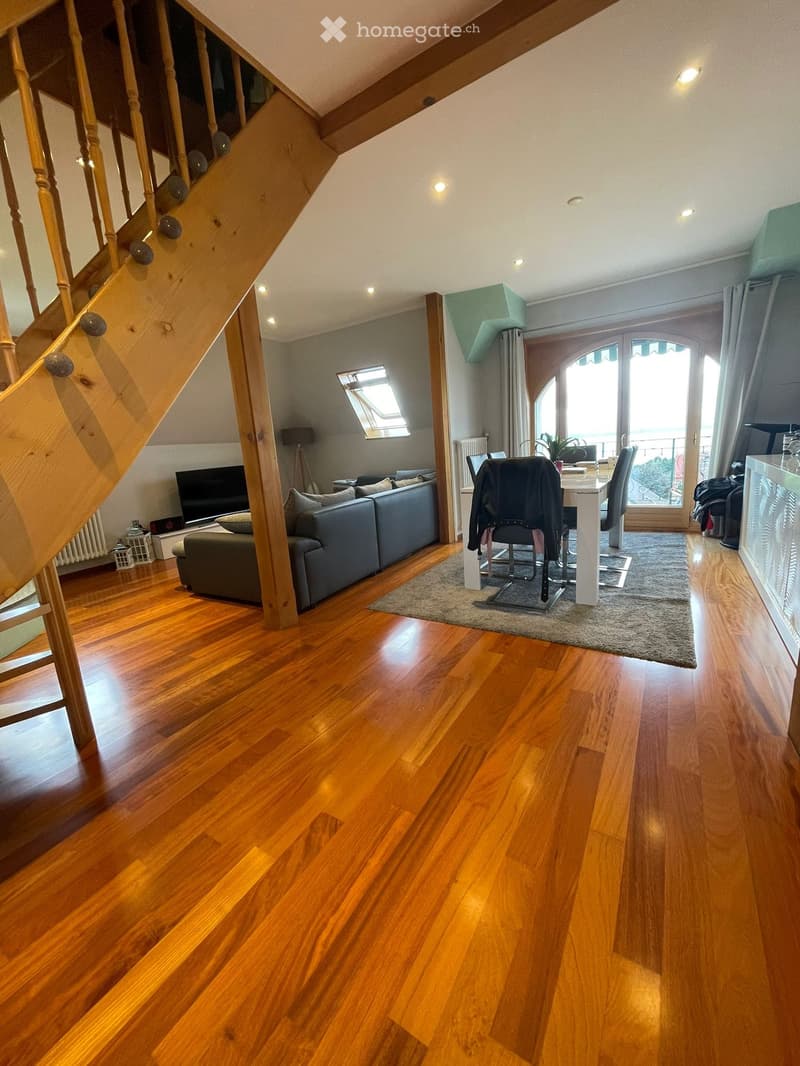Open-plan living and dining area with wooden flooring, a grey sofa, a dining table with chairs, and a television. The space features large windows and a staircase leading to an upper level.