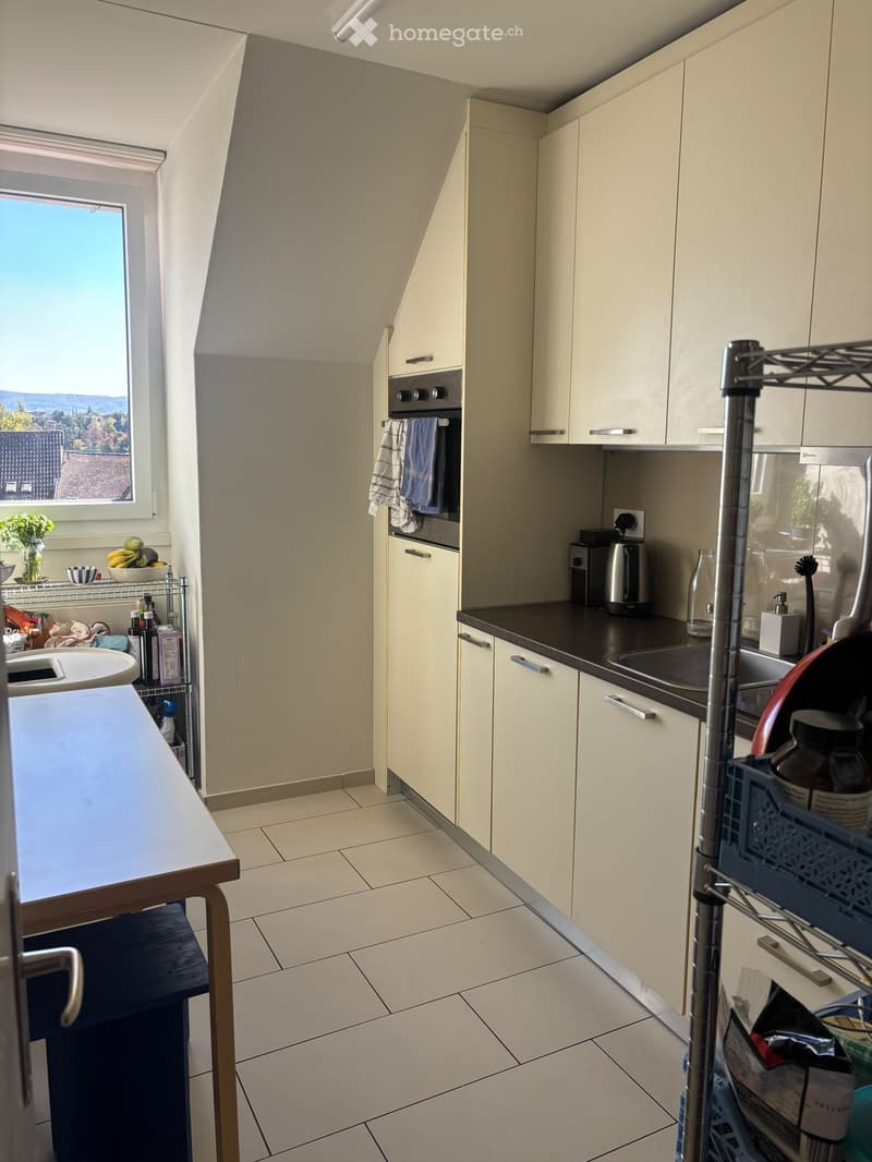 Modern kitchen with light-colored cabinets, a sink, an oven, and a countertop. There is a small table with a vase of flowers and a bowl of fruit on a metal shelf, along with various kitchen items and utensils.