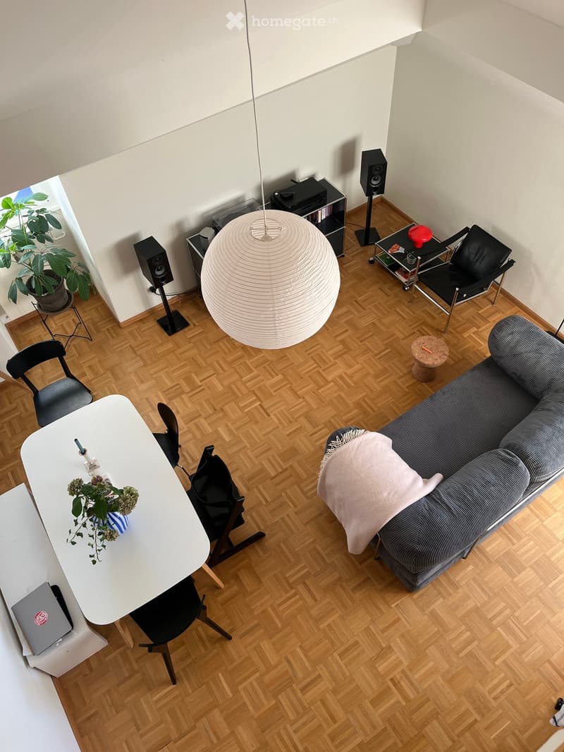 A modern living room with a grey sofa, white coffee table, black chairs, a laptop, a large hanging lamp, speakers, and a plant in the corner.