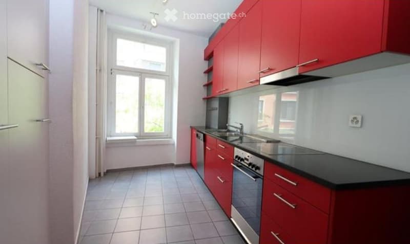 Modern kitchen with red cabinets, black countertops, stainless steel appliances, and a large window.