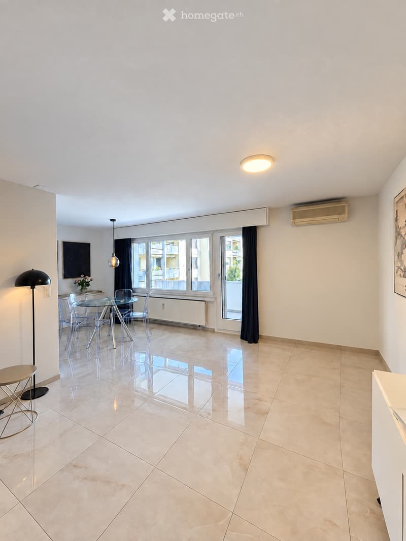 Bright, modern dining area with a glass dining table, four chairs, a floor lamp, and a small round stool. The room features large windows with black curtains, a polished tile floor, and a ceiling light fixture.
