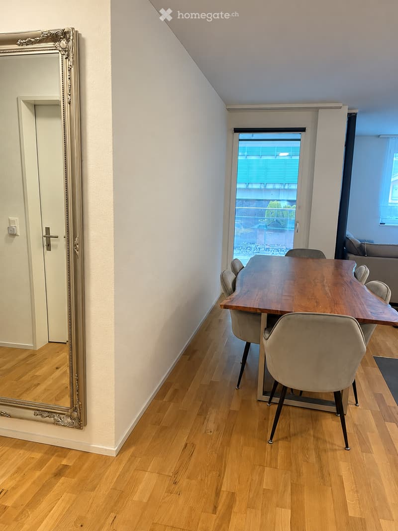 Dining area with a wooden dining table and chairs, large mirror on the wall, and a view of a balcony through a glass door.