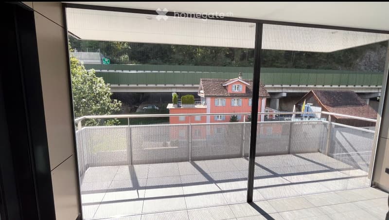 Balcony with modern glass sliding doors, metal railing, tiled floor, and a view of a cityscape with buildings and a bridge.